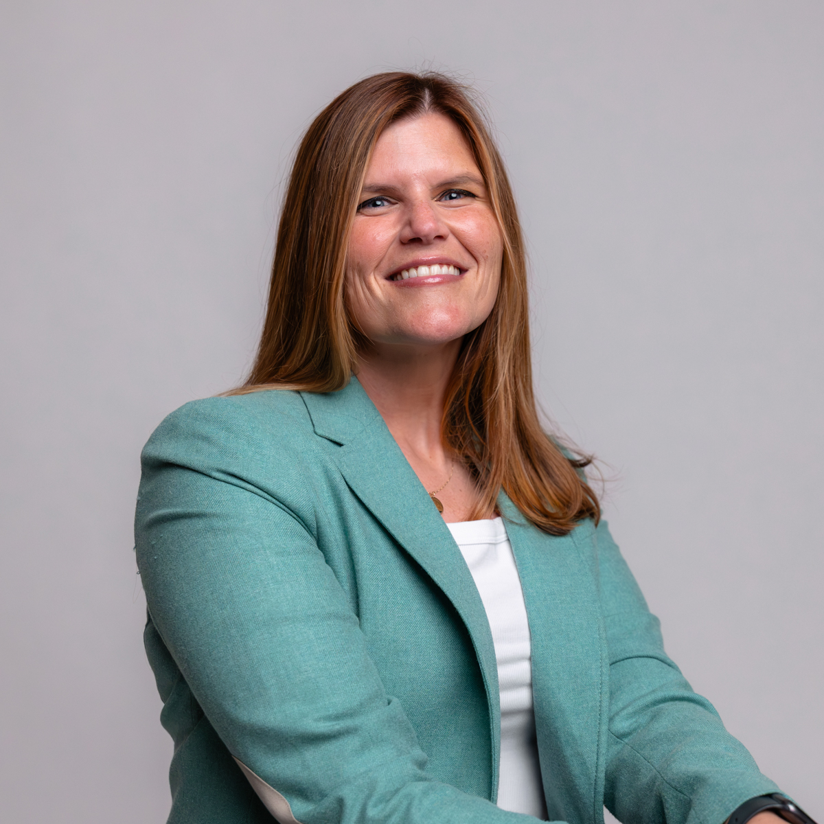 A woman with long brown hair, wearing a light teal blazer and white top, smiles while sitting against a plain light gray background.