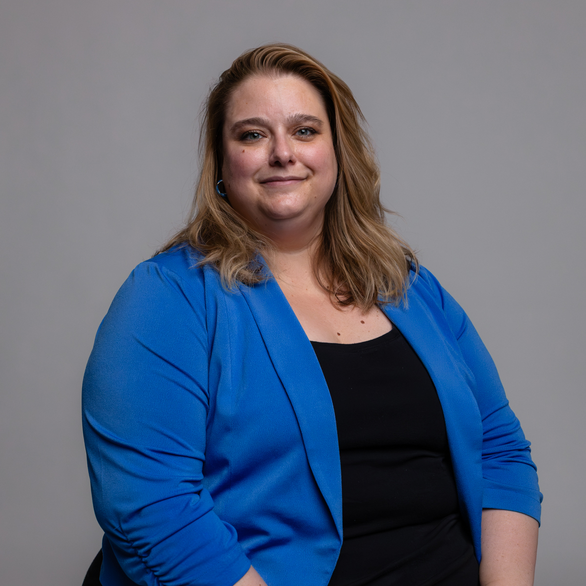 A woman with long, light brown hair wearing a blue blazer over a black top sits and smiles at the camera against a plain gray background.
