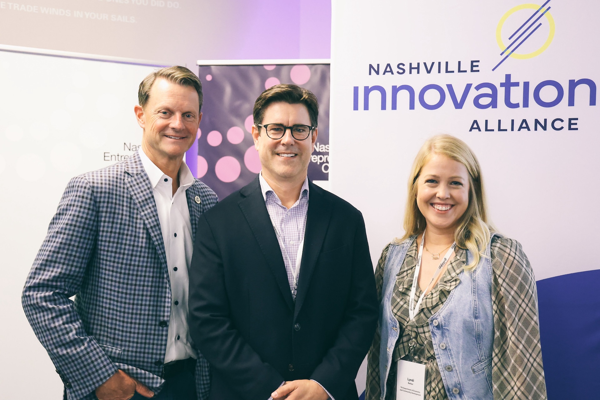 Three professionally dressed people smiling at a Nashville Innovation Alliance event, standing in front of banners with the organization’s logo and branding.