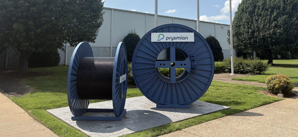 Two large blue cable reels, one full and one empty, are displayed outdoors on a paved platform in front of a building. One reel has a “Prysmian” sign attached. Green grass, bushes, and trees surround the area.