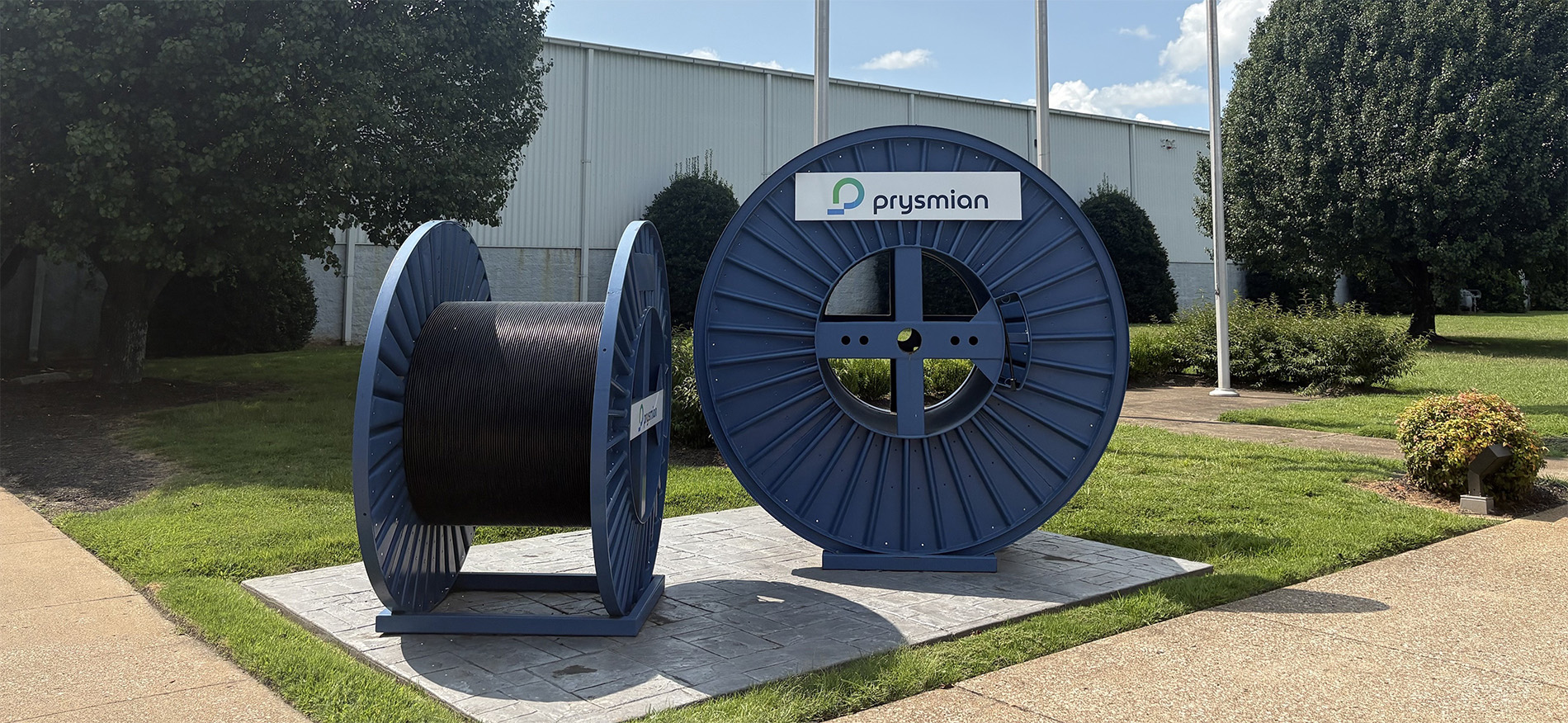 Two large blue cable reels, one full and one empty, are displayed outdoors on a paved platform in front of a building. One reel has a “Prysmian” sign attached. Green grass, bushes, and trees surround the area.