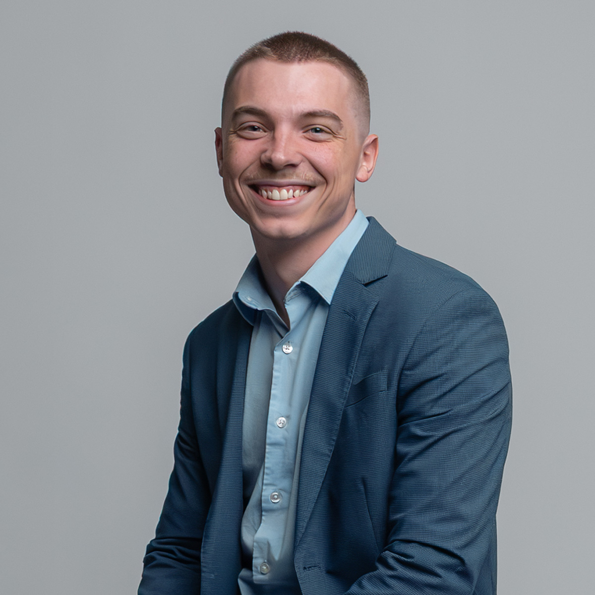 A young man with short hair, wearing a blue suit and light blue shirt, sits smiling against a plain light gray background.