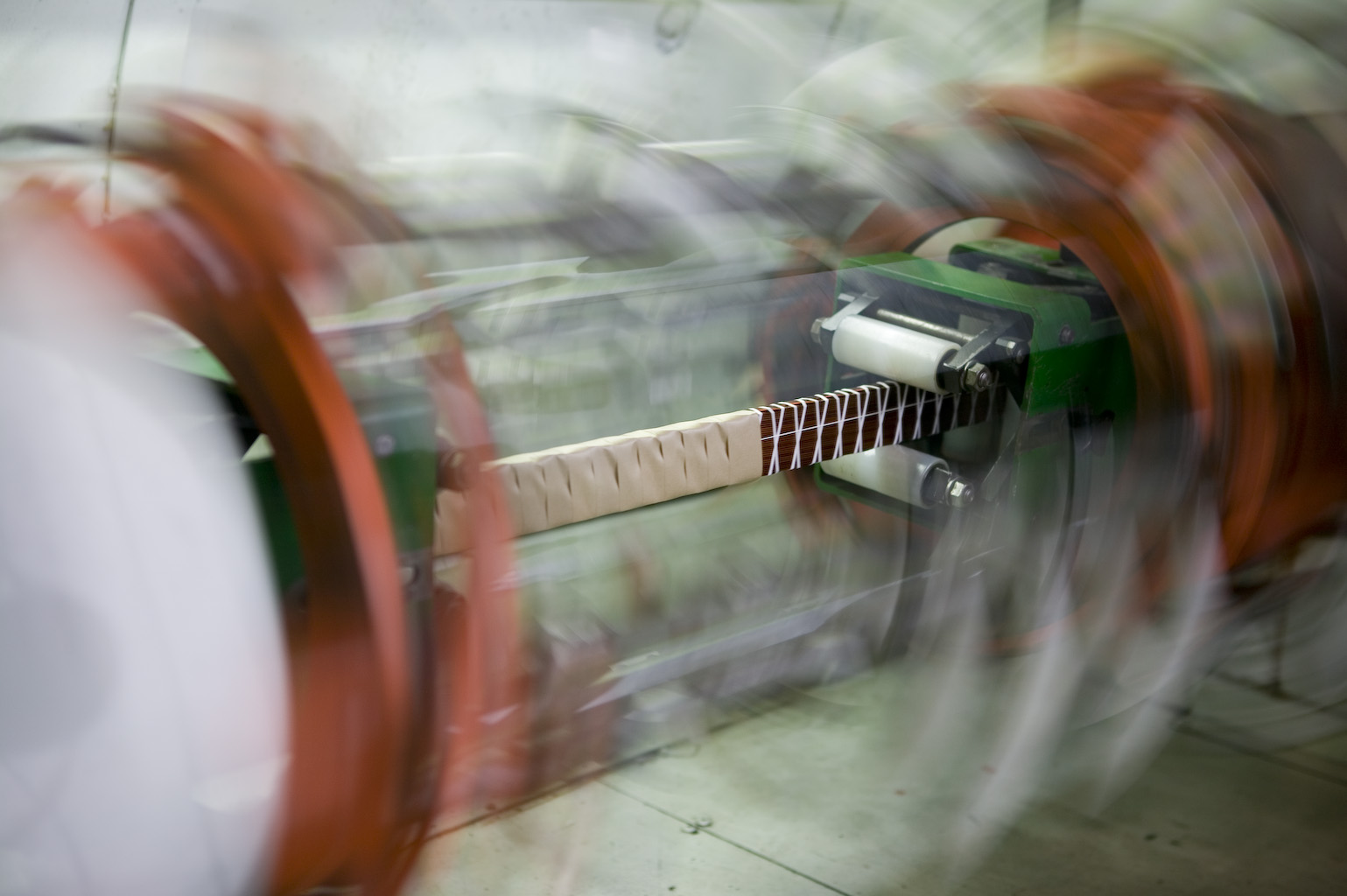 A machine with spinning orange parts twists or braids a beige cable or tube, creating a patterned surface. The motion blur emphasizes the fast movement of the equipment.