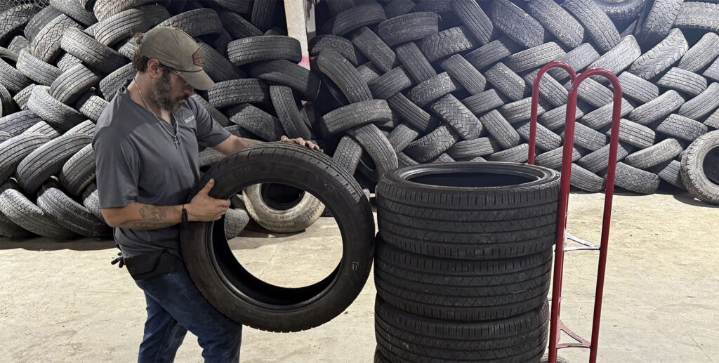 A person holding a tire next to a stack of tires, with a large pile of used tires in the background and a red dolly nearby in a warehouse-like setting.