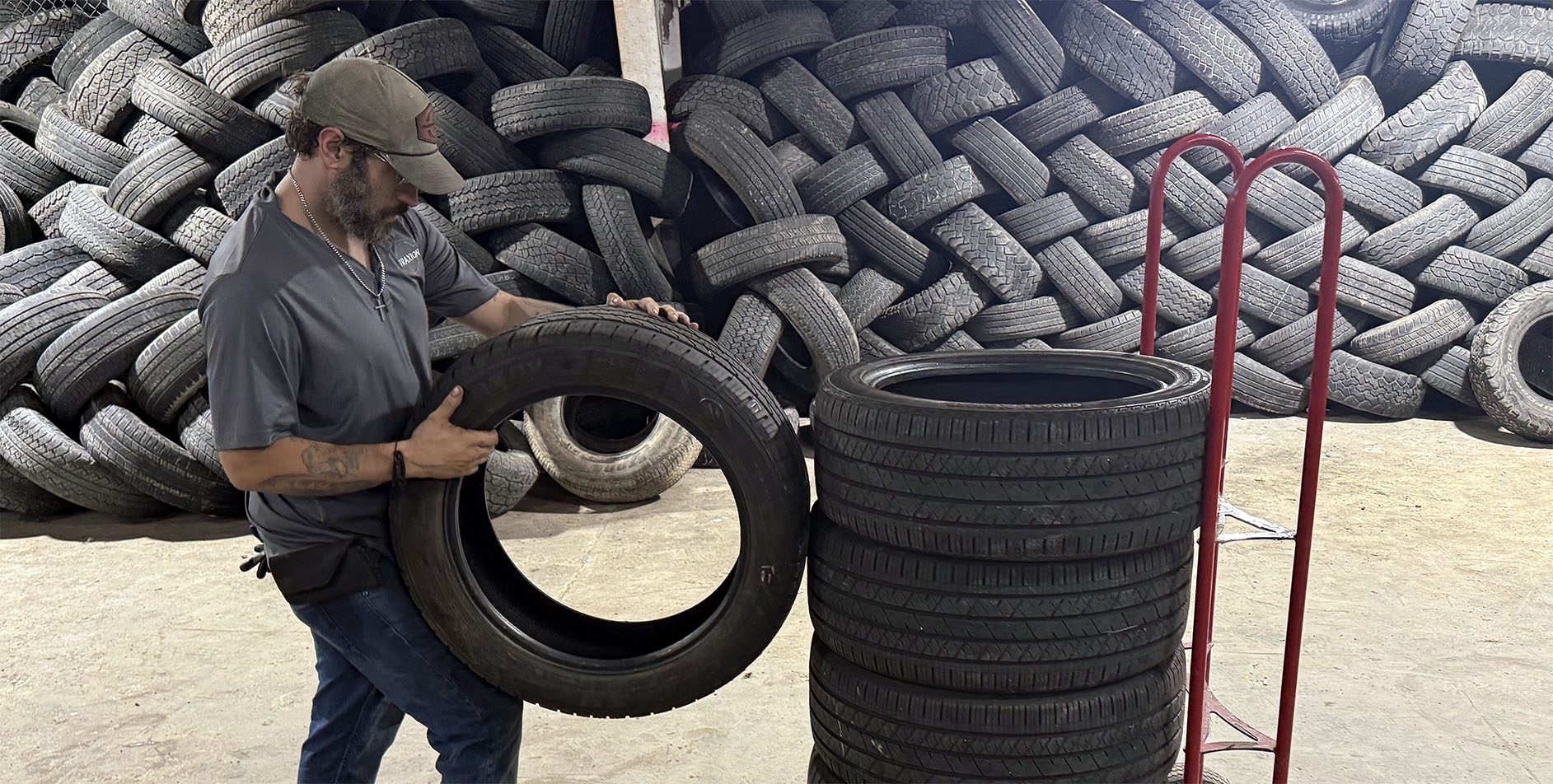 A person holding a tire next to a stack of tires, with a large pile of used tires in the background and a red dolly nearby in a warehouse-like setting.