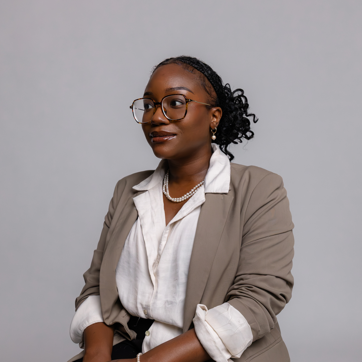 A woman wearing glasses, a light blazer, and a white shirt sits against a plain, light background. She has braided hair pulled back and is accessorized with pearl earrings and a matching necklace.