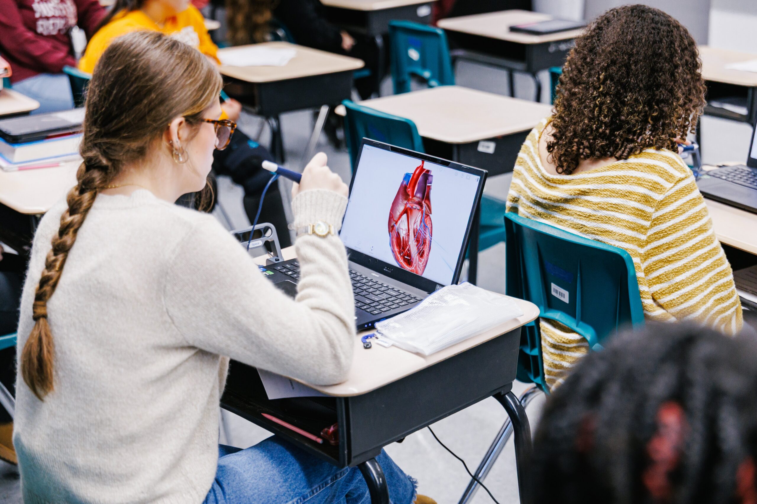 A student with a long braid uses a laptop displaying a heart diagram in a classroom setting. Other students sit at desks nearby, some using laptops.