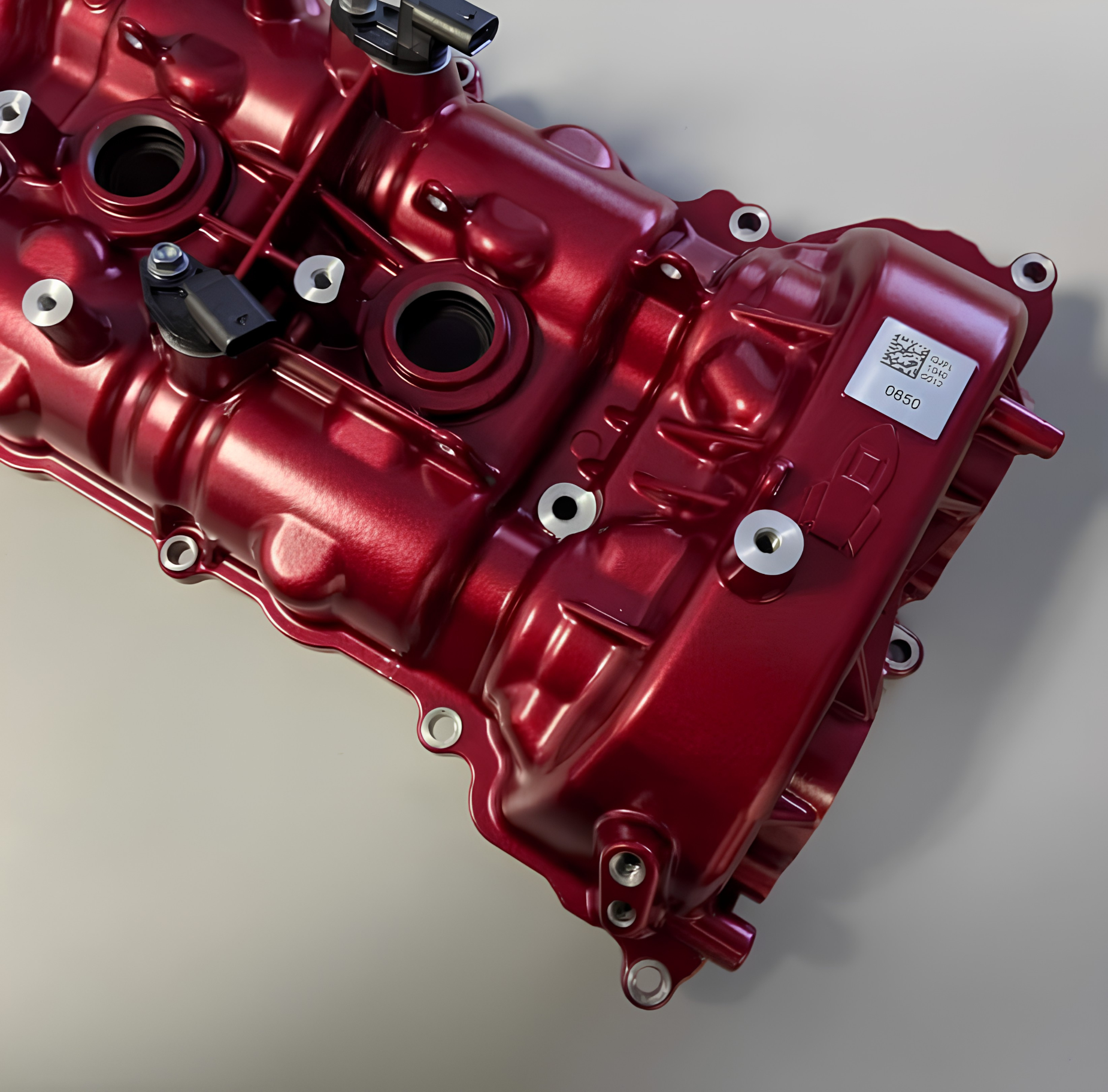 A metallic red engine valve cover with several bolt holes and a white label rests on a light-colored surface.