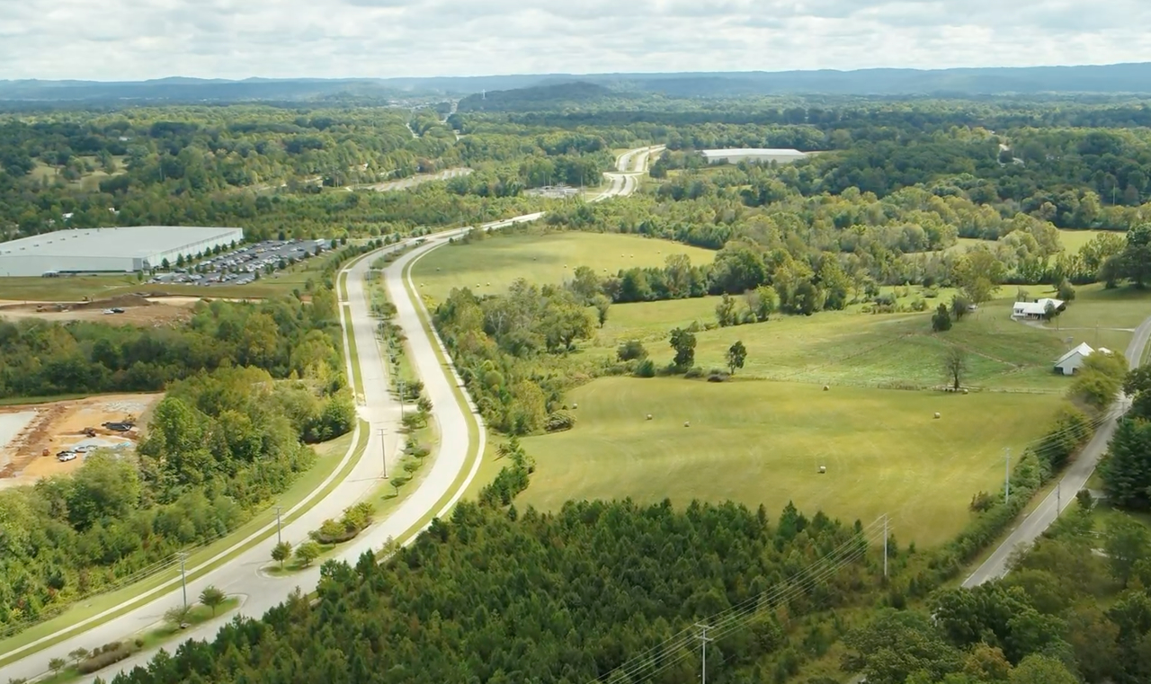 A winding road curves through green fields and wooded areas under a partly cloudy sky, with buildings, parking lots, and distant hills visible in the background.