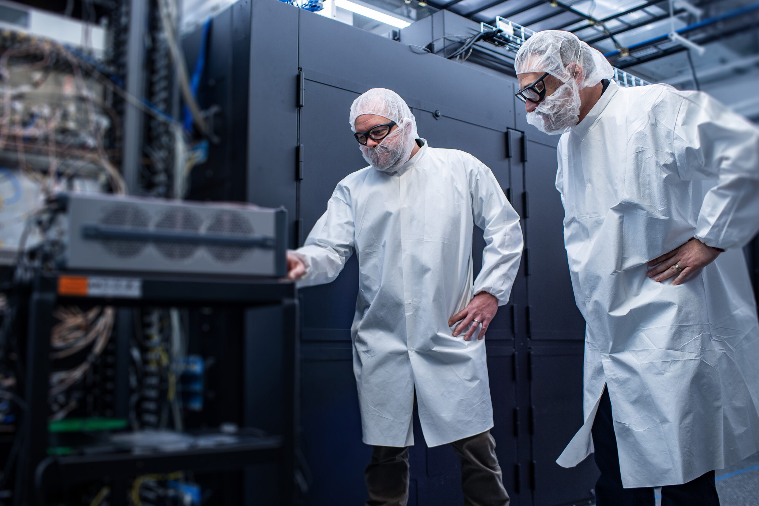 Two people in lab coats, hairnets, and safety glasses stand in a high-tech lab, examining equipment. One points at a machine, while the other observes. Computer servers and cables are visible in the background.