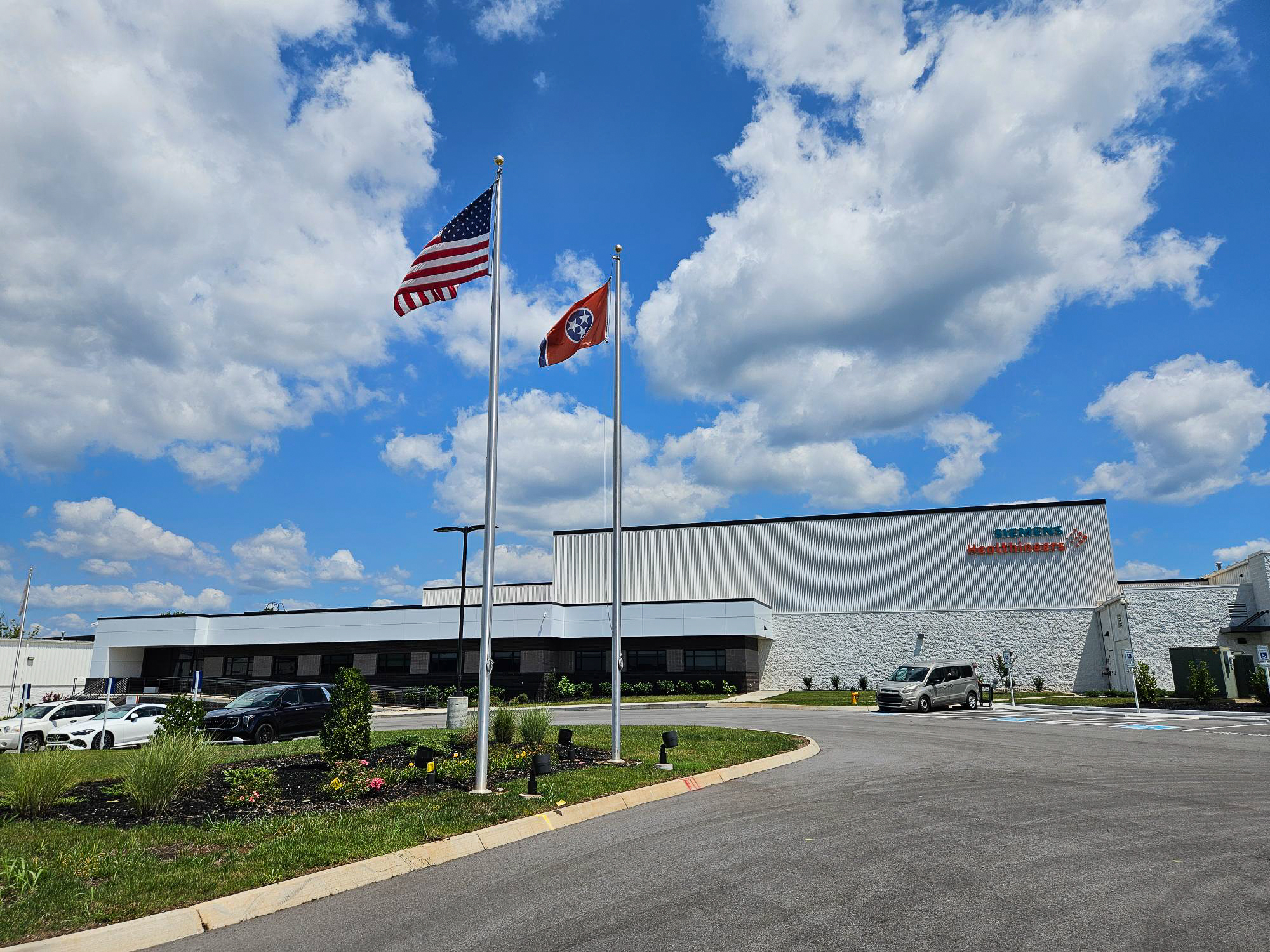 Two flagpoles with the US and Tennessee flags stand in front of a Siemens Healthineers building under a bright blue sky with scattered clouds. Cars are parked along the curved driveway.