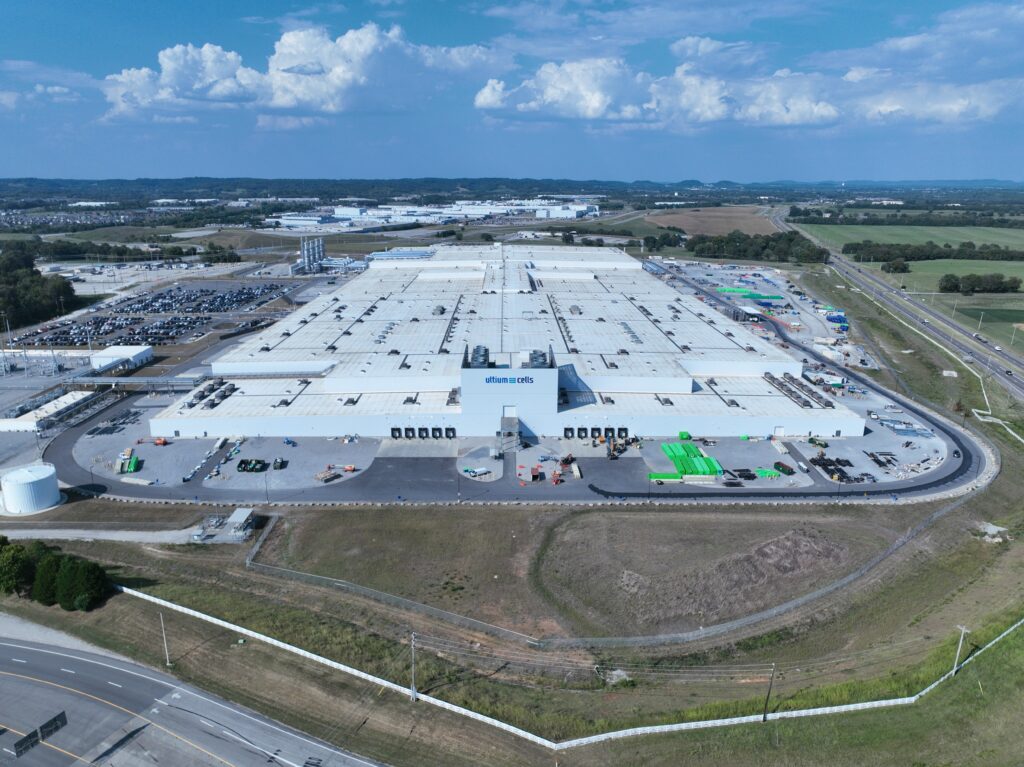 A large industrial warehouse complex with white roofs, surrounded by parking lots, roads, green fields, and a partly cloudy sky, viewed from above.