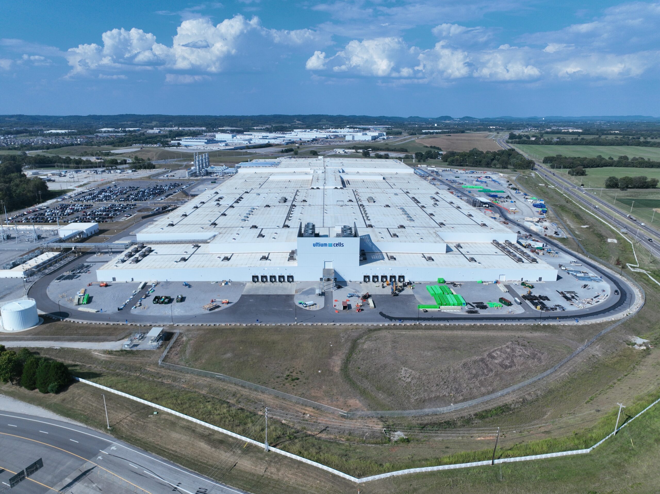 A large industrial warehouse complex with white roofs, surrounded by parking lots, roads, green fields, and a partly cloudy sky, viewed from above.