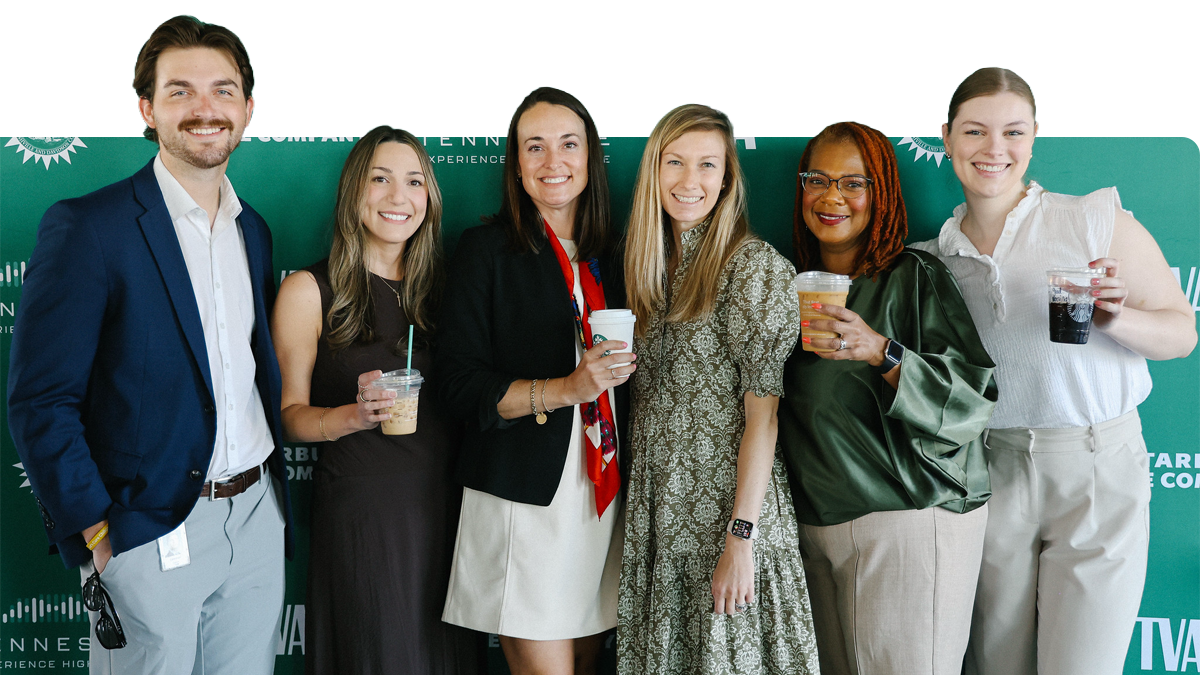 Six people stand smiling in front of a green event backdrop, each holding a drink. They are dressed in business or business-casual attire and appear to be enjoying a professional gathering.