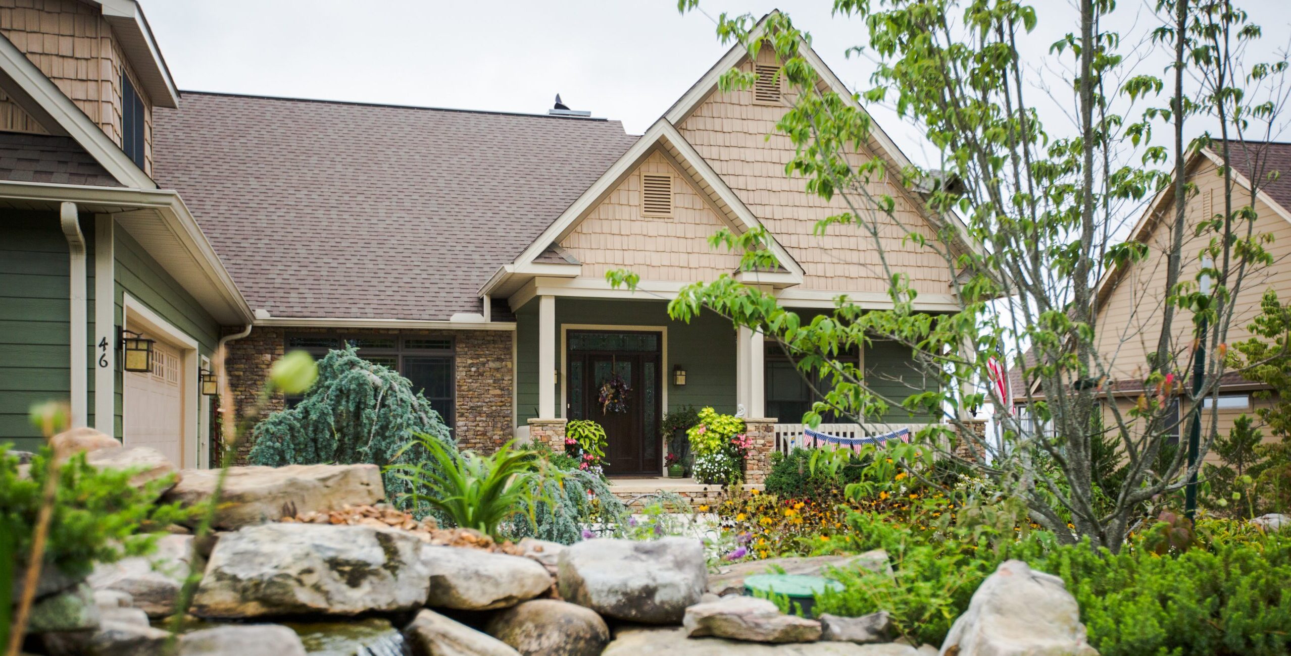 A single-story house with green siding and a stone entryway, viewed from behind a rocky garden pond with a small waterfall.