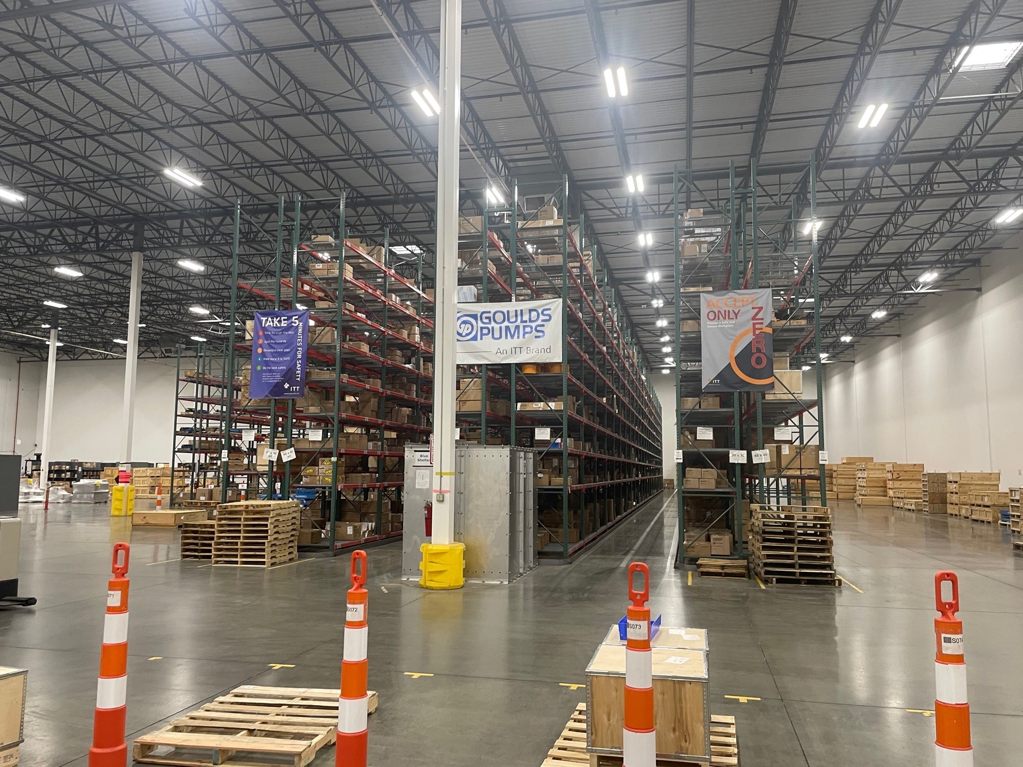A warehouse interior with high metal shelving units, aisles of stored boxes, and safety signage for Goulds Pumps and workplace safety protocols.