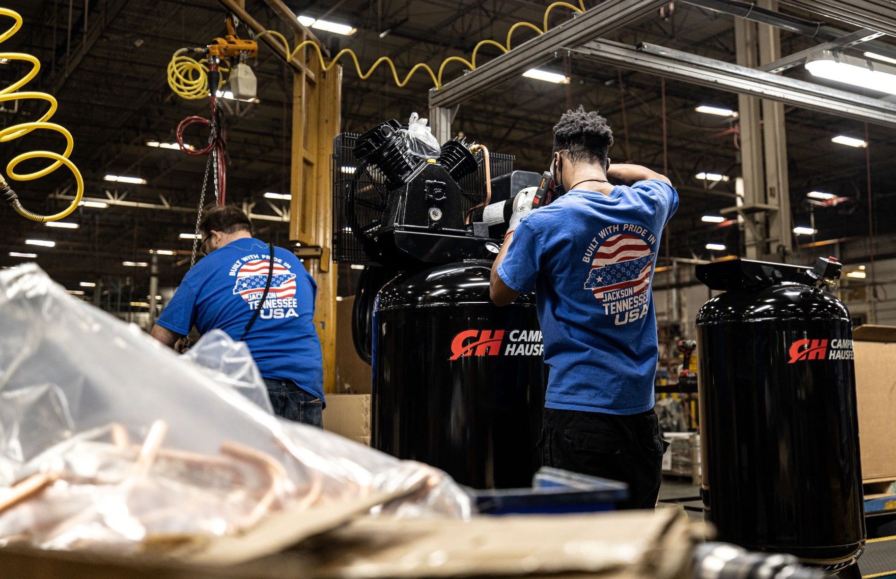 Two workers in blue company shirts assemble black air compressors on a factory floor.