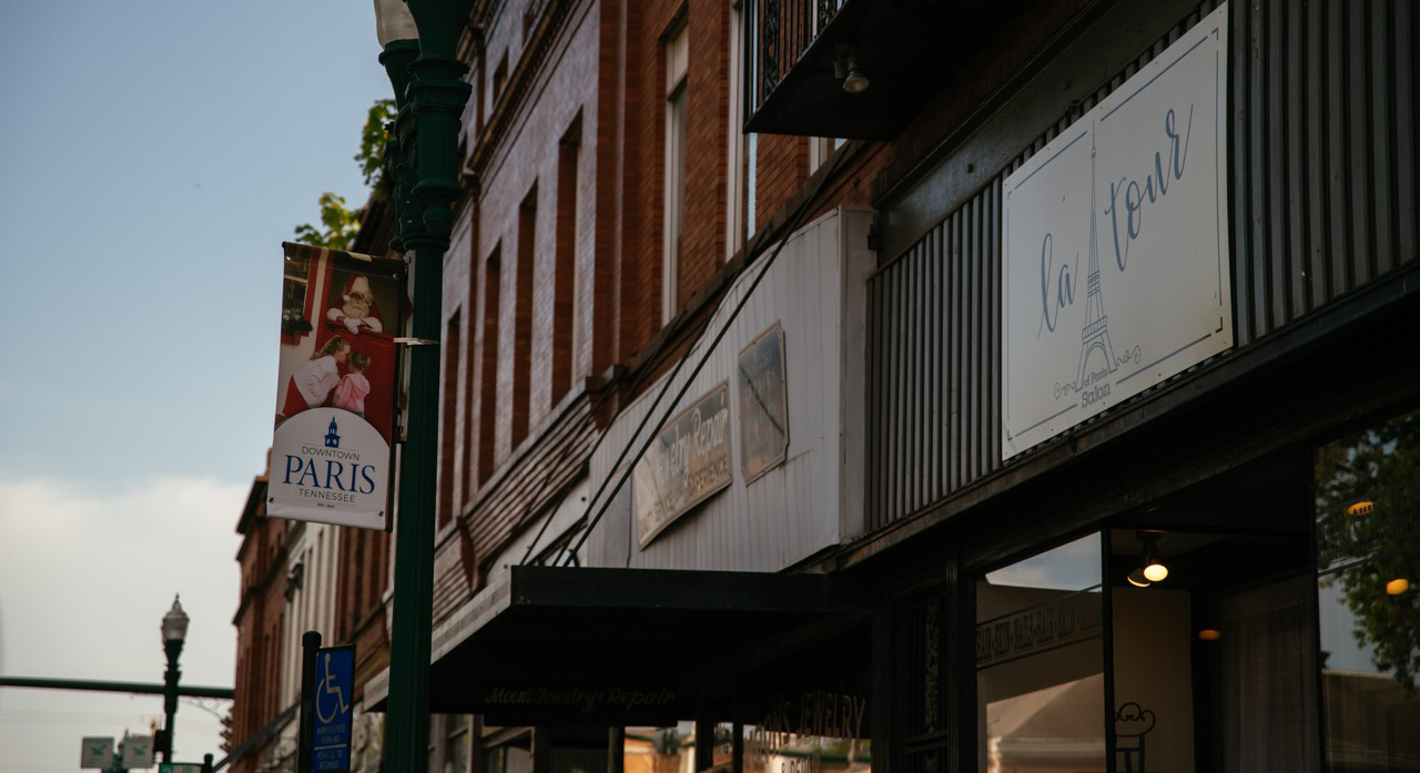 A view of a downtown street in Paris, Tennessee, featuring a banner for the city and signs for a local jewelry repair shop and a salon named La Tour.