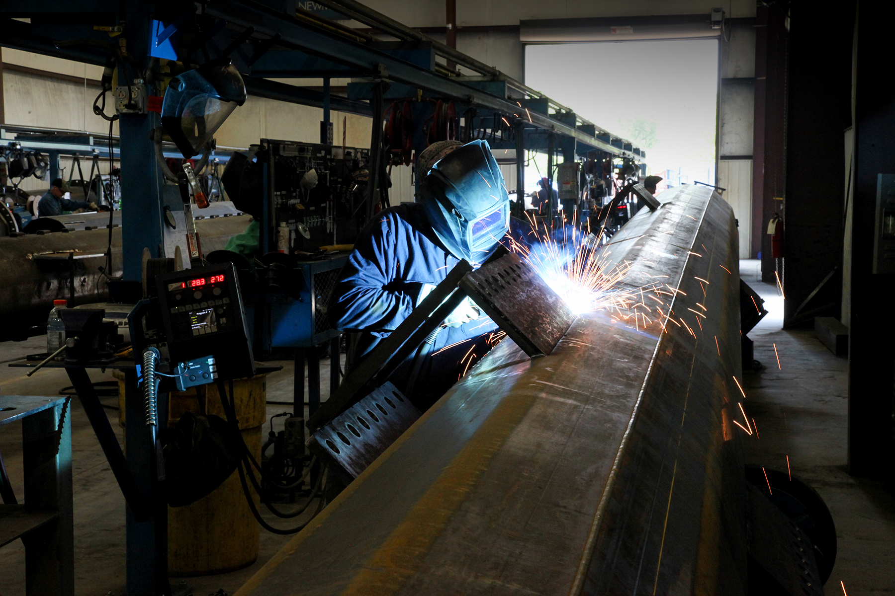 A worker wearing a welding helmet welds a large steel beam, sending sparks into the air in a workshop.