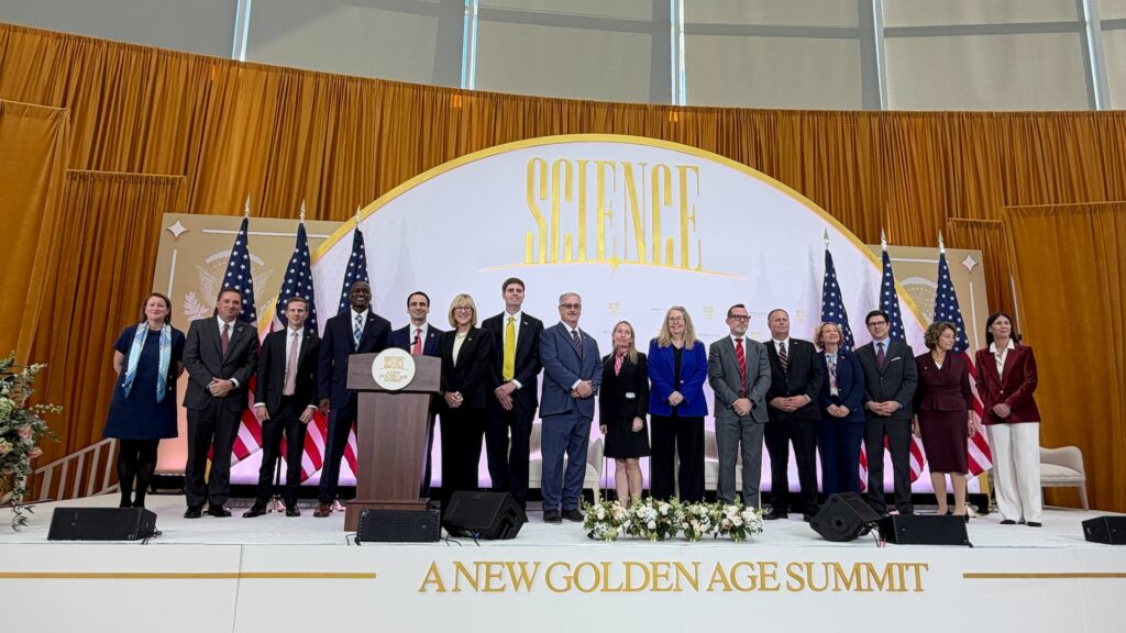 A group of sixteen people stand on a stage in front of a “Science: A New Golden Age Summit” backdrop, with several people positioned behind a wooden podium.